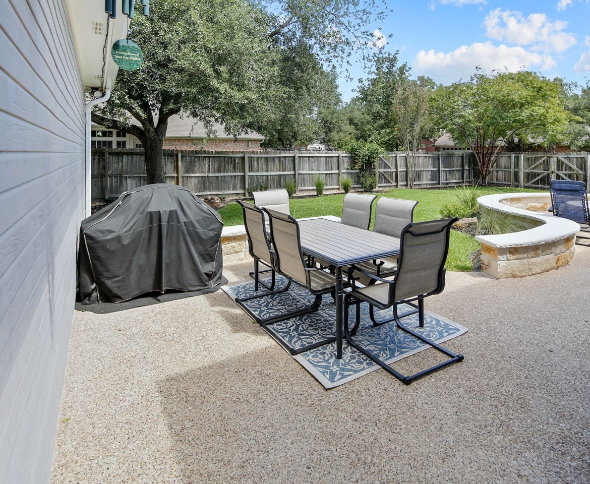 A patio with table and chairs, grill and trees.